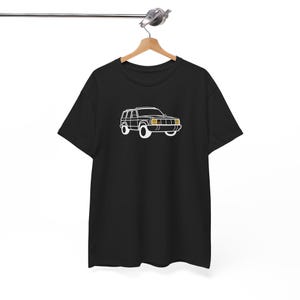 Puede incluir: Camiseta negra con un contorno blanco de un SUV clásico. Los faros del vehículo están resaltados en amarillo. La camiseta está colgada de una percha de madera.