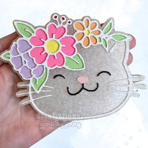 May include: A glittery silver cat-shaped decoration with a floral crown. The flowers are pink, orange, and purple with green leaves. The cat has a pink nose, black eyes, and whiskers. The text "Scentsational Scents by Jenny" is at the bottom.