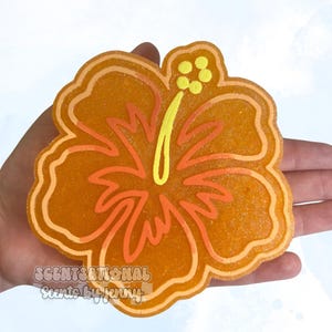 May include: Orange hibiscus flower-shaped item with a yellow and orange design. The item has a glittery texture and the text "SCENTSATIONAL Scents by Jenny" is visible. The flower's petals and center are detailed with varying shades of orange and yellow.