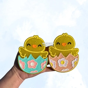 May include: Two yellow chick-shaped decorations, each emerging from a pastel eggshell. The eggshells are decorated with floral patterns in pink and white. The chick's beaks are orange, and they have smiling faces. The background is a light blue.