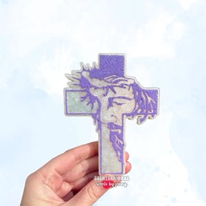 May include: A decorative cross featuring a detailed image of Jesus with a crown of thorns, rendered in purple glitter. The cross is held against a soft, watercolor-style background. The text "SCENTSATIONAL Scents by jenny" is visible.