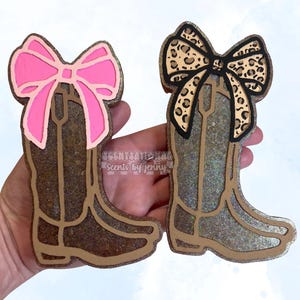 May include: Two decorative cowboy boot ornaments. One boot has a pink bow, the other a leopard print bow. Both boots are brown with glitter accents and a gold outline. The text "Scentsational Scents by Jenny" is visible.
