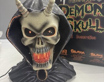 May include: A Fiberoptic Demon Skull decoration with a black hooded cloak. The skull is off-white with glowing red eyes and a red illuminated mouth. The horns are light beige. The box behind the skull reads "Fiberoptic Demon Skull Decoration."