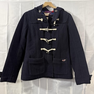 May include: A navy blue duffel coat with a hood and toggle closures. The coat has two front pockets and a small logo on the lower right side. The coat is hanging on a wooden hanger.