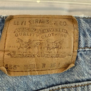 May include: Close-up of a light brown Levi Strauss & Co. label on blue denim jeans. The label features text including "ORIGINAL QUALITY CLOTHING" and a graphic of two horses. The jeans are Relaxed Fit, with a waist size of 36 inches and a length of 30 inches.