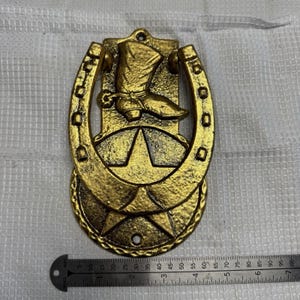 May include: A gold-colored metal door knocker featuring a horseshoe shape, a cowboy boot, and a star motif. A metal ruler placed at the bottom shows the object measures approximately 6.5 inches in total height from top to bottom edge.