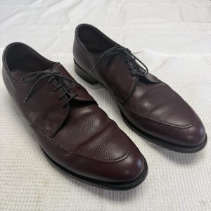May include: A pair of dark burgundy leather derby shoes featuring a textured, pebbled grain finish, a rounded toe box with a stitched apron seam, and thin dark laces threaded through four eyelets on each side of the central opening.