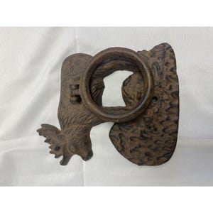 May include: A rustic, cast iron door knocker in a brown finish. The door knocker features a stylized moose design with a ring for knocking. The moose has antlers and a textured surface.