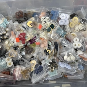 May include: A collection of various buttons in small, clear plastic bags. The buttons are in a variety of colors, including white, blue, red, and black. Some buttons have decorative designs. The buttons are of different sizes and shapes.