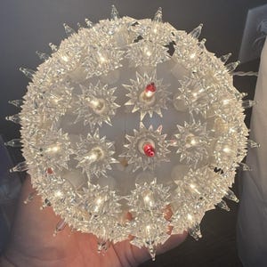 May include: A clear, spherical Christmas light decoration with a starburst design. The sphere is covered in clear, spiky plastic pieces, with small, warm-toned lights and a few red lights. The overall effect is a festive, glowing orb.