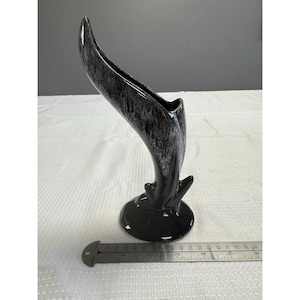 May include: A tall, black and gray ceramic vase with a unique, curved design. The vase has a speckled pattern and sits on a round base. The vase is approximately 10 inches tall.