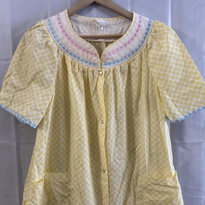May include: Yellow vintage robe with a small square pattern. The robe has short sleeves with blue trim, a decorative collar with pink and blue accents, and two pockets. It buttons down the front.