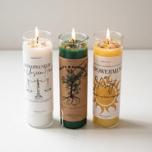 May include: Three cylindrical glass jar candles with lit wicks. The left candle is white with a scale graphic and text reading Entrepreneur. The center candle is green with a tree graphic and text reading Unity & Opportunity. The right candle is yellow with a sun graphic and text reading Empowerment & Clarity.