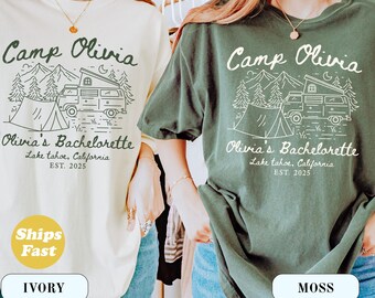 May include: Ivory and moss green t-shirts with a camping-themed graphic. The design includes the text "Camp Olivia," a camper van, tent, trees, and the text "Olivia's Bachelorette, Lake Tahoe, California, EST. 2025."