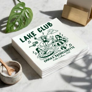 Puede incluir: Una pila de servilletas de papel cuadradas blancas con un estampado gráfico en verde oscuro. El diseño incluye el texto LAKE CLUB, EMMA'S BACHELORETTE y Lake Tahoe, 2026, junto con ilustraciones de tres personas en traje de baño.