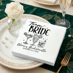 May include: White paper napkins with the text "Team Bride" in black script, along with illustrations of cocktails. The napkins are personalized with "Olivia's Bachelorette MARBELLA 2026". A white rose in a small glass vase is on the left.