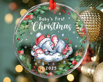 May include: A clear Christmas bauble with a red ribbon. The bauble features a sleeping baby elephant wearing a Santa hat on a cloud, surrounded by greenery and the text "Baby's First Christmas 2025".