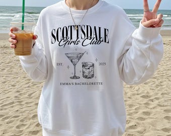 Può includere: Felpa bianca con la scritta "SCOTTSDALE Girls Club" e una grafica di cocktail. Sotto la grafica c'è la scritta "EST. 2025 EMMA'S BACHELORETTE". La felpa è indossata su una spiaggia sabbiosa.