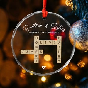 May include: A clear, round Christmas ornament with the words "Brother & Sisters" and "FOREVER LINKED TOGETHER" in elegant script. The ornament features a crossword puzzle design with the names JAMES, OLIVIA, and BAILEY. A red ribbon is attached for hanging.