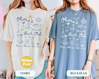 May include: Two oversized t-shirts, one cream and one blue, with a beach-themed graphic. The design includes the text "Olivia's Bach Club, Miami, Florida, Est. 2025" with illustrations of cocktails, umbrellas, and seashells. The cream shirt has a "Ships Fast" sticker.