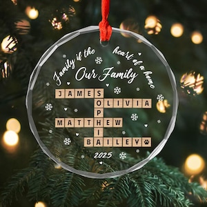 May include: A clear, round Christmas ornament with the text "Family if the heart of a home, Our Family." Names are spelled out in wooden letter tiles. The year 2025 is at the bottom. A red ribbon is attached.