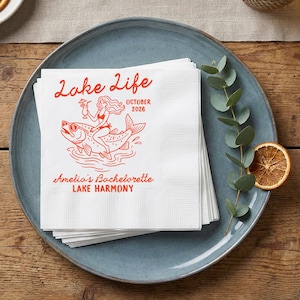 May include: A stack of white paper napkins featuring a red graphic print. The design includes the text Lake Life, October 2026, Amelia's Bachelorette, and Lake Harmony, with an illustration of a person riding a fish while holding a drink.