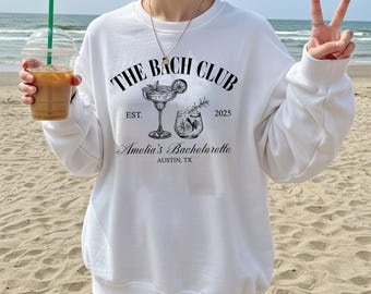 Può includere: Felpa bianca con la scritta "THE BACH CLUB" e illustrazioni di cocktail. Sotto, il testo recita "Amelia's Bachelorette, Austin, TX, EST. 2025". La felpa ha una vestibilità ampia.