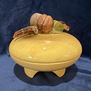 Puede incluir: Un cuenco redondo de cerámica amarilla con tapa y acabado moteado, apoyado sobre tres patas cortas. La tapa presenta un grupo decorativo de frutas marrones y hojas verdes esculpidas en tres dimensiones sobre la superficie superior.