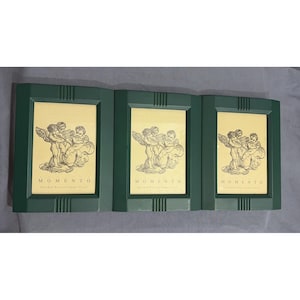 Peut inclure: Trois cadres photo rectangulaires vert foncé disposés côte à côte. Chaque cadre contient un insert couleur crème avec un dessin au trait noir représentant trois chérubins et le texte "MOMENTO" imprimé en lettres capitales noires.