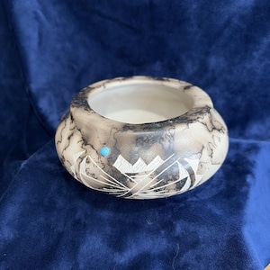 May include: A round ceramic bowl with a smooth, cream-colored surface featuring dark, web-like smoke patterns. The exterior is decorated with carved geometric white lines and a single small, circular turquoise-colored inlay on the side.