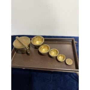 May include: A set of brass measuring cups and a small circular weight marked with the number 12, arranged on a rectangular brown tray. The set includes one lidded cup and four open cups of varying sizes, all featuring a polished gold-tone finish.