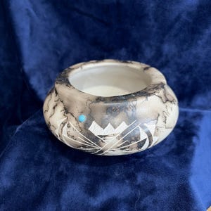May include: A round ceramic bowl featuring a cream-colored base with dark, smoky black horsehair-style markings. The exterior is decorated with carved white geometric patterns and a single small, round, bright blue turquoise-colored inlay.