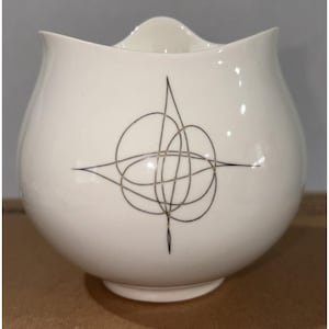 May include: Cream-colored ceramic bowl with a unique, abstract black and gold design. The bowl has a rounded body and a slightly scalloped rim. The design is a series of intersecting lines and shapes, adding a touch of mid-century modern style.