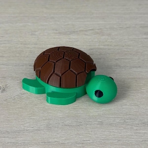 May include: A 3D-printed turtle figurine. The turtle has a brown, textured shell and a green body, including the head, flippers, and base. The turtle's head has a black eye. The figurine is sitting on a light-colored wooden surface.