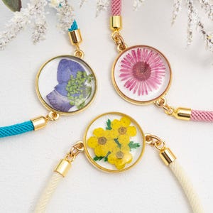 Puede incluir: Tres pulseras con colgantes circulares dorados que contienen flores prensadas. Una pulsera tiene un cordón turquesa, otra un cordón rosa y la tercera un cordón crema. Los colgantes presentan una flor morada, una flor rosa y flores amarillas.