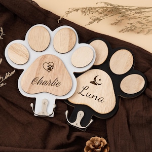 Pode incluir: Dois porta-chaves em forma de pata, um branco e outro preto, com almofadas de madeira. O suporte branco tem o nome "Charlie" e um coração com pegada de pata. O suporte preto tem o nome "Luna" e um desenho de lua e estrela. Ambos têm ganchos prateados.