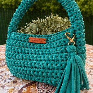 May include: A teal crochet handbag featuring a rounded handle, a textured stitch pattern, and a gold-toned metal clasp with a matching teal fabric tassel. A rectangular brown leather-like tag with the text Susia Jouj is attached to the front.