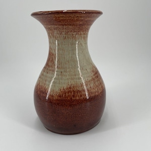 May include: A handcrafted ceramic vase with a flared top and a rounded body. The vase features a two-tone color scheme, with a deep reddish-brown base that fades into a lighter tan color towards the top. The vase has a glossy finish.