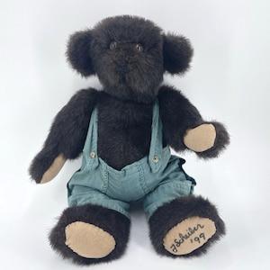 May include: A dark brown teddy bear wearing teal overalls. The bear has tan paw pads and ears. The bear is signed "J. Scheiber '99" on one foot. The bear is sitting against a white background.