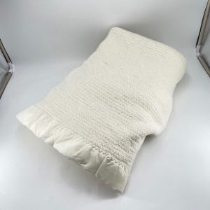 May include: Ivory-colored knitted blanket with a satin trim. The blanket has a textured, woven appearance and a soft, plush satin border. The blanket is folded, showcasing the texture and the contrasting satin edge.