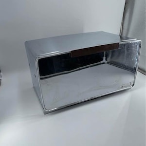May include: A shiny, chrome-finished bread box with a rectangular shape. It features a dark brown wooden handle and a hinged door. The box is designed with clean lines and a minimalist aesthetic, suitable for kitchen storage.