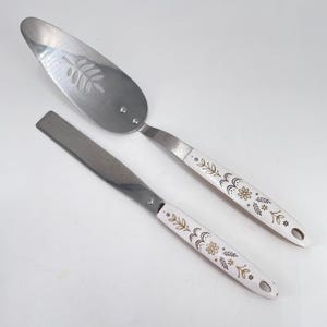 May include: A stainless steel cake server and a spatula. The server has a slotted blade with a leaf design. Both utensils have white handles decorated with gold and brown floral patterns. The handles have a hole at the end for hanging.