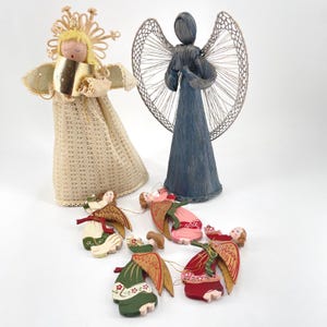 May include: A collection of six angel figurines including two standing figures and four smaller hanging ornaments. One standing angel wears a cream lace dress with a gold halo, while the other is blue with wire wings. Four painted wooden angels feature colorful robes.