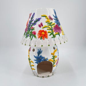 May include: A white ceramic lamp with a floral design. The lamp features a mushroom-shaped base with a cut-out opening and a lampshade decorated with colorful flowers and butterflies. The lamp has a white picket fence design around the lampshade.
