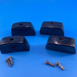 May include: Four black, trapezoidal-shaped knobs with a smooth, glossy finish. Each knob features a single centered hole on the top surface. Four small, metallic, threaded screws are positioned separately below the knobs on the surface.