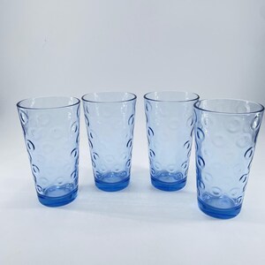 May include: A set of four light blue drinking glasses, each featuring a repeating embossed circular pattern across the exterior surface. The glasses are cylindrical in shape with a smooth rim and a flat base, showcasing a consistent translucent color.