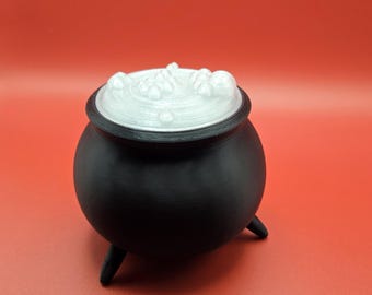 May include: A black cauldron with three legs and a white, bubbly substance on top. The cauldron is set against a solid red background. This decorative item is perfect for Halloween or fantasy-themed decor.