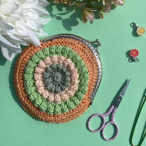 Puede incluir: Una cartera de monedas de ganchillo con un diseño circular en verde, rosa y naranja. La cartera tiene un marco de metal plateado y está sobre una superficie verde.