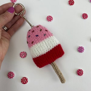May include: A pink, white and red knitted ice cream cone keyring with a brown string. The keyring is decorated with colourful sprinkles.
