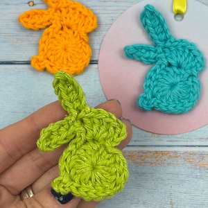May include: Three crocheted bunny shapes in different colors: orange, teal, and lime green. The bunnies are all the same size and have a simple design.